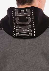 Kapuzensweatshirt mit schwarzer Kapuze, die mit silbernen Nieten und Schriftzügen versehen ist. Der Hauptkörper ist dunkelgrau und hat eine glatte Textur.