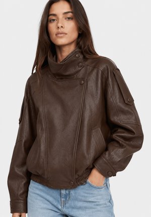 Femme aux longs cheveux bruns portant une veste en cuir marron foncé avec un col haut et un jean bleu clair sur un fond uni.