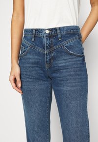 Mörkblå denimjeans med hög midja, femfickorsdesign, frontknäppning och framträdande sömnadsdetaljer på fickorna.