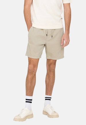 Beige cotton shorts with an elastic waistband, drawstring, and side pockets. Worn with white sneakers and striped socks.