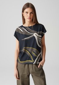 Bluzka z krótkim rękawem w kolorze granatowym z abstrakcyjnymi wzorami w beżu, bieli i zieleni. Wykonana z gładkiego materiału, o luźnym kroju i okrągłym dekolcie.