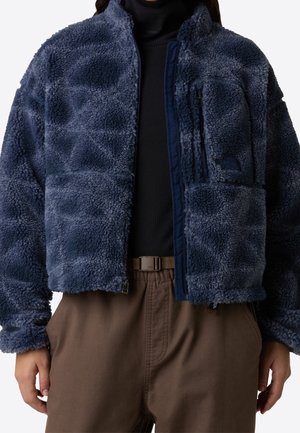 Personne portant une veste polaire bleu marine sur un col roulé noir et un pantalon marron avec une ceinture marron, debout les mains détendues sur les côtés.