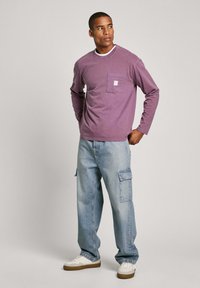 Camiseta de algodón morada de manga larga con un bolsillo en el pecho. Combinada con unos jeans de corte suelto en azul claro que tienen bolsillos tipo cargo, y zapatillas blancas.