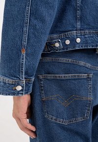 Jeansjacka med en strukturerad passform, mörkblå färg, silverfärgade tryckknappar och synliga sömdetaljer. Har en ficka med ett zigzag-mönster.