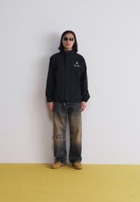 Jachetă pullover neagră cu fermoar frontal și logo, asociată cu blugi blue deteriorați și pantofi bej. Fundal galben.