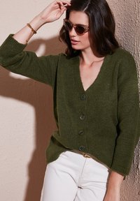 Groene gebreide cardigan met een V-hals, knopen aan de voorkant en driekwart mouwen, tegen een gestructureerde lichte achtergrond.