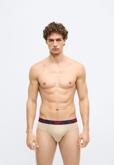 Cuecas masculinas bege com uma cintura elástica vermelha e azul-marinho apresentando um logótipo. O tecido é suave e ajustado, destacando os contornos do corpo.