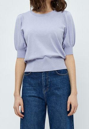 Femme portant un pull court à manches bouffantes de couleur violet clair, rentré dans un jean en denim bleu foncé taille haute, debout devant un fond uni.