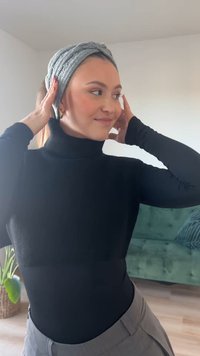 Črna turtleneck majica z dolgimi rokavi, kombinirana s sivo teksturirano trakcem za lase. Model stoji ob zeleni sedežni garnituri v dobro osvetljenem prostoru.