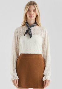 Donna bionda che indossa una blusa bianca in pizzo a maniche lunghe, una gonna in suede marrone e una sciarpa nera con motivo, in piedi contro uno sfondo grigio chiaro.