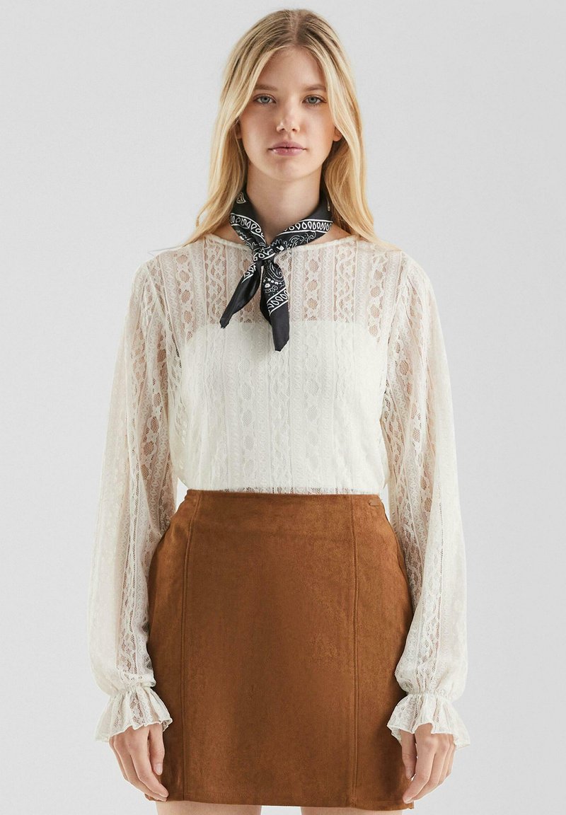 Donna bionda che indossa una blusa bianca in pizzo a maniche lunghe, una gonna in suede marrone e una sciarpa nera con motivo, in piedi contro uno sfondo grigio chiaro.