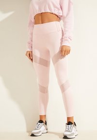Leggings deportivos de color rosa claro con paneles de malla, diseño de cintura alta y una textura suave. Combinados con zapatillas negras y blancas.