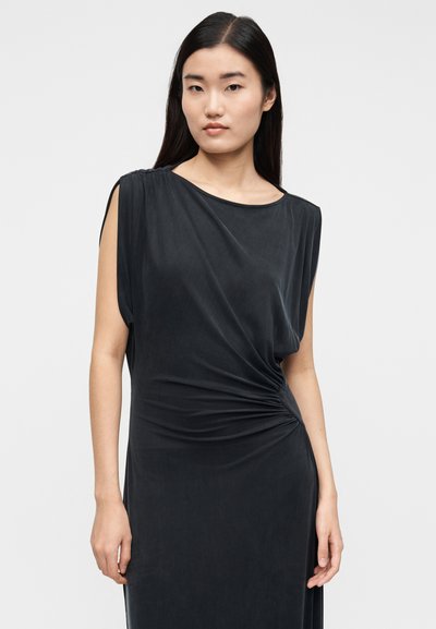 Woman with long dark hair wearing a black sleeveless dress with side slit detail, standing against a plain light background.