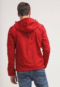 Giacca leggera rossa con cappuccio, caratterizzata da una chiusura con zip e cinghie regolabili. Il tessuto appare liscio con una texture minima.