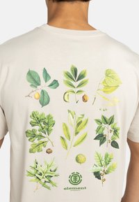 Cremefarbenes T-Shirt mit botanischen Illustrationen verschiedener Blätter und Nüsse in grünen und gelben Tönen, mit einem runden Logo am unteren Rand.