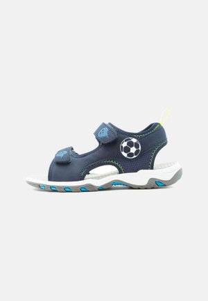 Marineblå børnesandal med to velcro-remme, fodbold-design, neon-gult trækkestrop og hvid sål med blå og grå accenter.