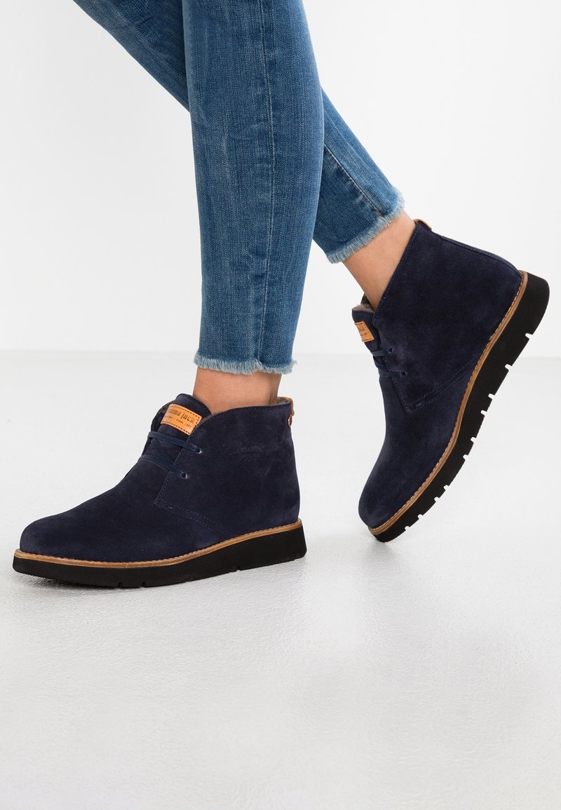 Bottines en daim bleu marine à bout rond, accents en cuir marron, et semelle texturée ; assorties à un jean bleu délavé.