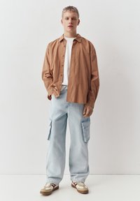 Młoda osoba stojąca, ubrana w brązową koszulę w kratę na białej koszulce, jasnoniebieskie spodnie cargo oraz beżowe trampki na jednolitym białym tle.