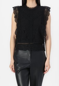 Top en dentelle noire avec bordure festonnée, mettant en avant des motifs floraux intricats et un col haut. Associé à un pantalon en cuir noir.