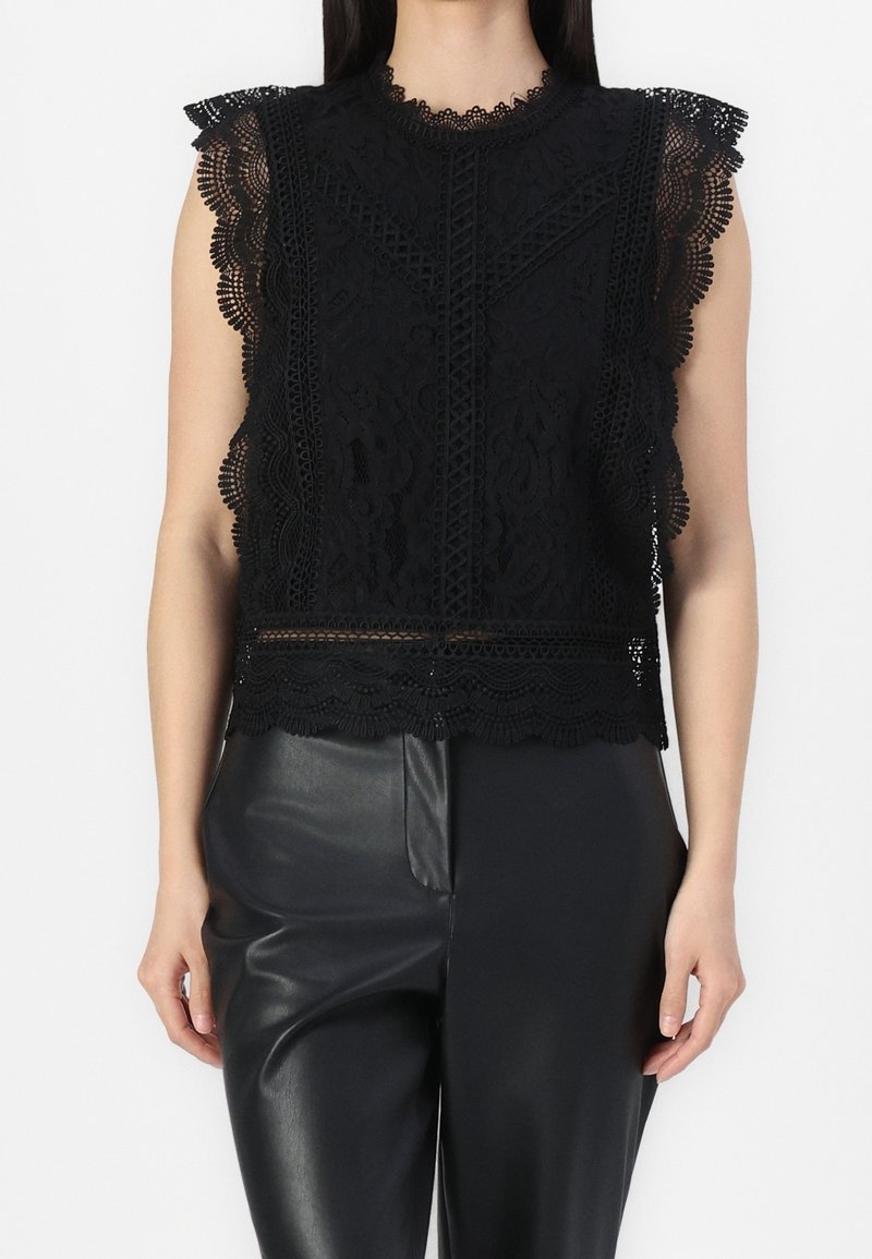 Top en dentelle noire avec bordure festonnée, mettant en avant des motifs floraux intricats et un col haut. Associé à un pantalon en cuir noir.