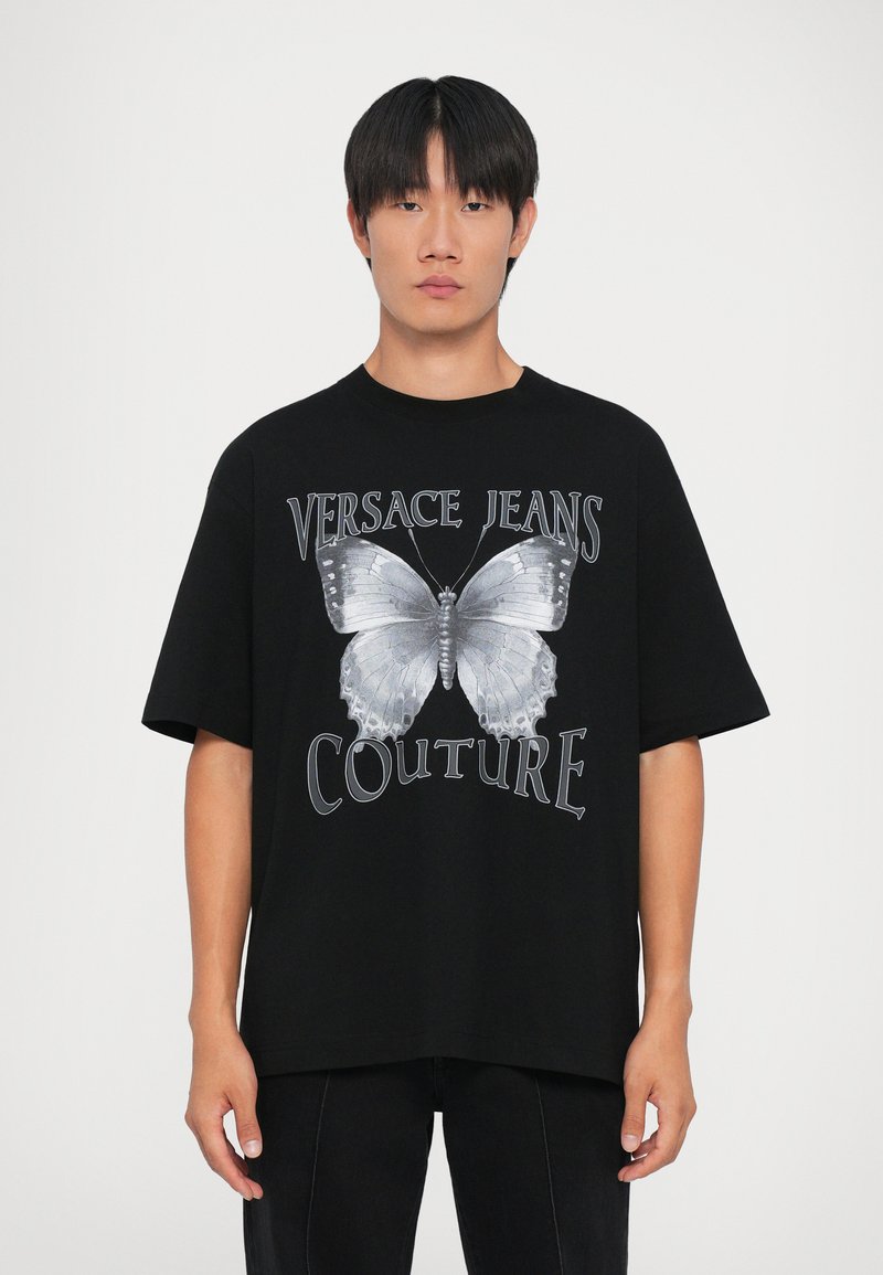 Νεαρός άνδρας που φοράει μαύρο T-shirt από Versace Jeans Couture με μεγάλη ασημένια γραφική πεταλούδα και κείμενο μπροστά, στέκεται μπροστά σε ουδέτερο φόντο.