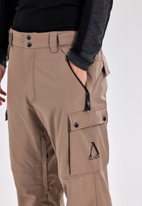 Tan cargobroeken met een gladde textuur, voorzien van een ritszak en decoratieve hardware. Logo op de bovenbeen, vlakke voorkant en riemlussen.