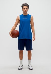 Νέος άνδρας στέκεται κρατώντας μπάλα του μπάσκετ στο δεξί του χέρι, φορώντας μπλε αμάνικο μπλουζάκι Under Armour, σκούρα μπλε σορτς, άσπρες κάλτσες και λευκά αθλητικά παπούτσια.