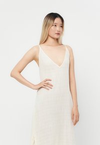 Rochie din in de culoare crem, cu decolteu adânc în formă de V și bretele subțiri. Siluetă dreaptă cu un material ușor texturat.