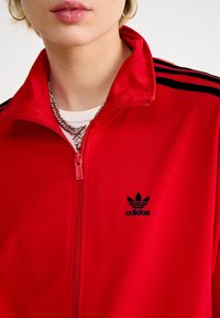 Punane adidasi tõmblukuga jakk, mis on valmistatud siledast kangast, mustade triipudega õlgadel ja logoga vasakul rinnal. Hõbedane tõmblukudetail.