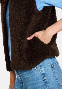 Brūns vilnas veste ar teksturētu, mīkstu virsmu, ar sānu kabatu un brīvu, atvērtu dizainu, valkāta virs zila džempera un džinsu.