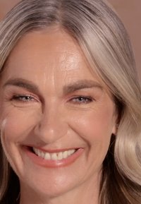 Primer plano de una persona con cabello rubio claro, piel suave y una sonrisa confiada. Presenta suaves arrugas alrededor de los ojos y la boca.