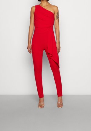 Vrouw die een rode jumpsuit met één schouder draagt met geplooide details aan één kant en zilveren sandaaltjes met bandjes, staand tegen een effen achtergrond.