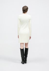 Knelang kremfarget ribbestrikket kjole med lange ermer, tettsittende silhuett, sidesplitter, brukt med teksturerte svarte knelange støvler.