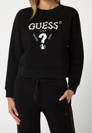 Kobieta ubrana w czarną bluzę z białym napisem „GUESS U.S.A.” i logo w formie znaku zapytania, do tego czarne spodnie z sznurkiem i bocznymi kieszeniami.