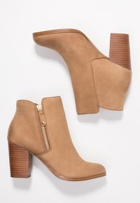 Stivali alla caviglia in pelle color tan caratterizzati da un tacco alto, punta arrotondata e chiusura con zip laterale in metallo dorato. Il tacco presenta delle scanalature orizzontali.