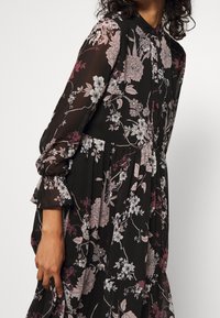 Woman wearing a black long-sleeve dress with pink and white floral patterns, buttoned front, and ruffled cuffs on a plain background.