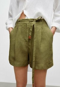 Olivgrønn linshorts med knyttet midje og oppbrettede ermer, med frontfolder og en liten lærdetalj på midjebåndet.