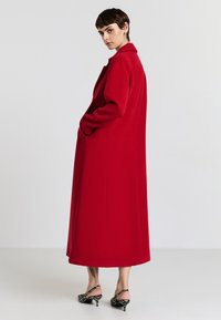 Roter, langer Mantel mit geradem Schnitt, großem Kragen und Seiten- taschen, aus einem strukturierten Stoff gefertigt. Kombiniert mit gestreiften offenen Schuhen mit Absätzen.