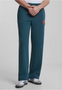 Pantalones de chándal color teal con cintura elástica, que presentan bolsillos laterales y un logo bordado en naranja. Fabricados con una tela suave y texturizada.