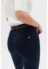 Jeans azul marino con un bolsillo trasero con un acento metálico, combinados con una camiseta blanca de canalé y un cinturón negro, vistos desde un ángulo lateral.