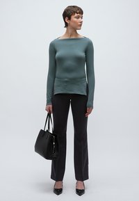 Hosszú ujjú, testhezálló, könnyű zöld felső sötét, egyenes szárú nadrággal párosítva. A modell egy fekete kézitáskát tart; a cipők hegyes orrúak.