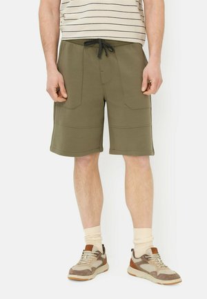 Mann trägt olivgrüne knielange Shorts mit Fronttaschen, gestreiftes Hemd, cremefarbene Socken und beige-braune Sneaker.