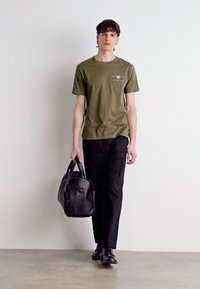 T-shirt verde oliva, pantalones cargo negros y bolsa de viaje de piel negra. Diseño sencillo, tejido texturizado y un logo sutil en la camiseta.