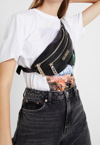 Sort faux læder bælteskede med tre lynlåslommer, accentueret af guldfarvet hardware, båret over en hvid grafisk T-shirt og sorte jeans.