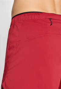 Red shorts with a textured fabric, featuring an elastic waistband and black drawstring. Side seam pockets are visible.