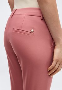 Κοντινό πλάνο σε ροζ παντελόνια με ραφή, με μια μικρή χρυσή καρφίτσα λογοτύπου πάνω από την πίσω δεξιά τσέπη, συνδυασμένα με ανοιχτό μπεζ τοπ.