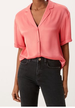 Femme portant une chemise corail rose à manches courtes et boutonnée, ample, rentrée dans un jean noir taille haute.