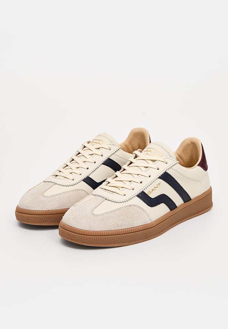 Sneaker mit einem Obermaterial aus cremefarbenem Leder, marineblauen gestreiften Akzenten und einer Wildlederspitze. Braune Gummisohle und Schnürsenkel in neutralen Farben.