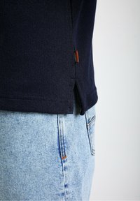 Temno modra majica s kratkimi rokavi z teksturirano tkanino, stranskimi razporka in majhno oznako blagovne znamke. Ujemajo se z svetlo modrim kavbojkam.