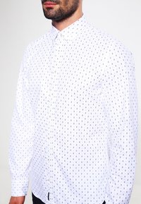 Weißes Hemd mit Knopfverschluss, das ein Rautenmuster in Dunkelblau aufweist. Lange Ärmel, klassischer Kragen und kleine Knöpfe unterstreichen das strukturierte Design.